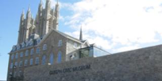 Exterior image of Guelph Civic Museum, a stone building set against a blue sky, with the Basilica of Our Lady spires in the background